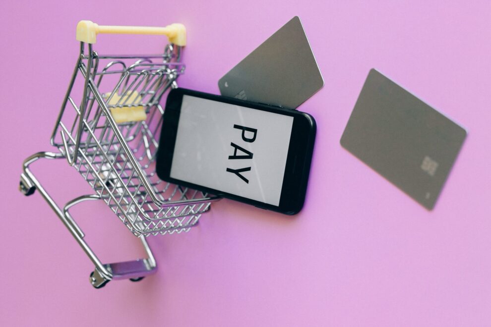 A mini shopping cart with a smartphone and credit cards falling out of it on a pink background.
